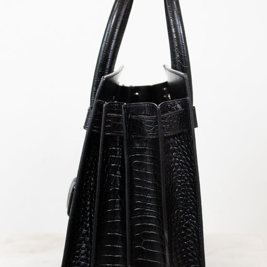 A black crocodile-embossed leather handbag shown from the back, featuring dual top handles and a structured silhouette, set against a plain white background.