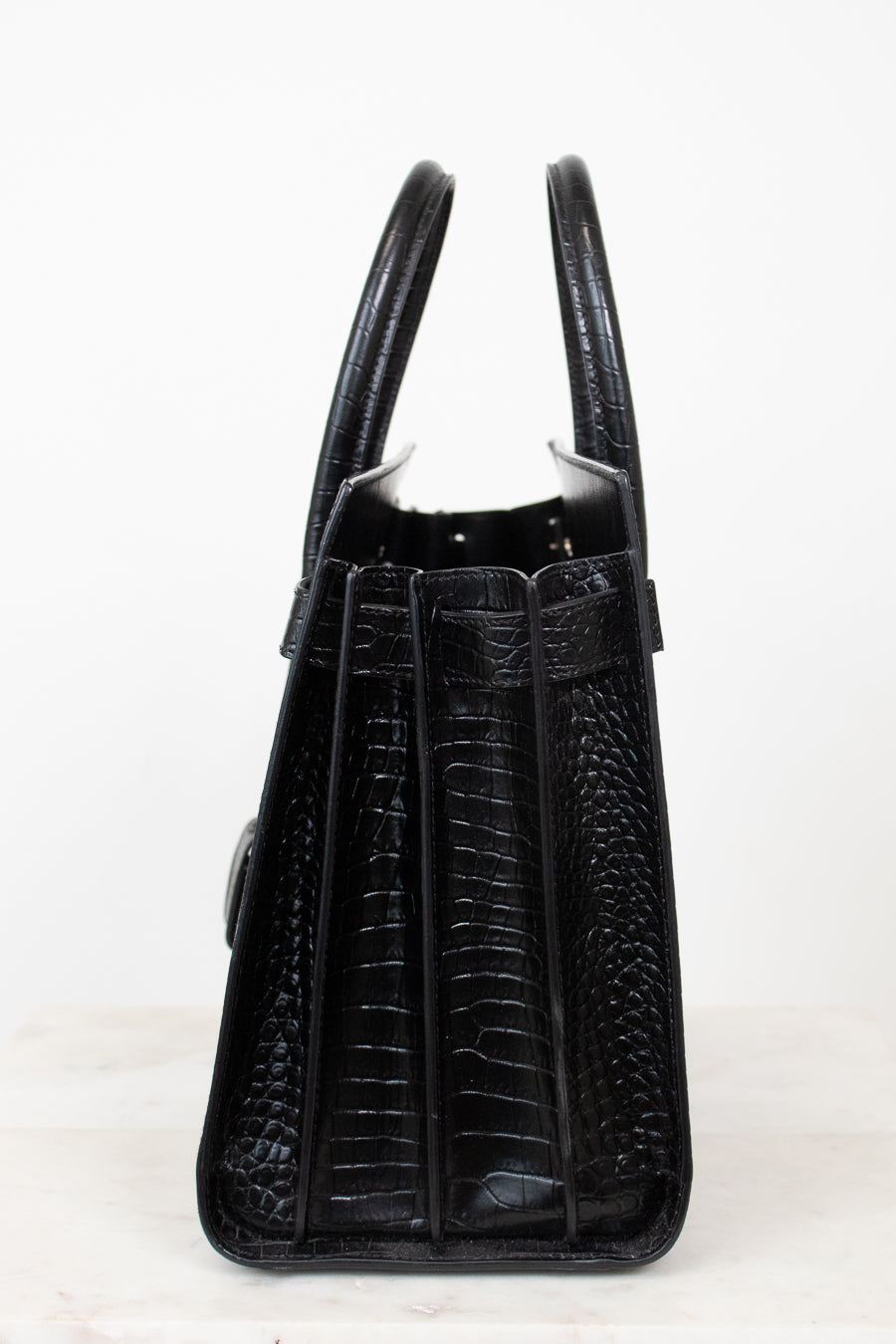 A black crocodile-embossed leather handbag shown from the back, featuring dual top handles and a structured silhouette, set against a plain white background.