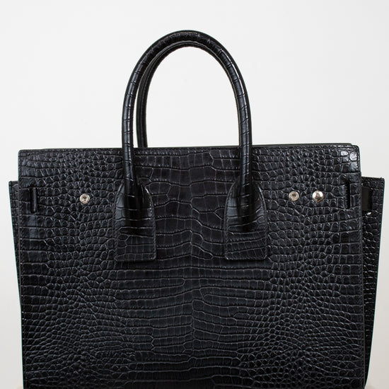 A black crocodile-embossed leather handbag with double top handles, displayed on a light marble surface against a plain white background.