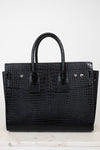 A black crocodile-embossed leather handbag with double top handles, displayed on a light marble surface against a plain white background.