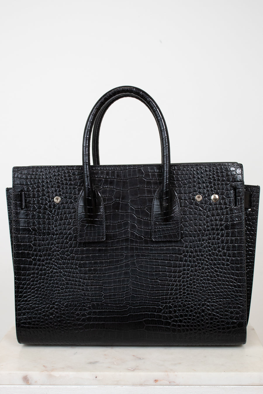 A black crocodile-embossed leather handbag with double top handles, displayed on a light marble surface against a plain white background.