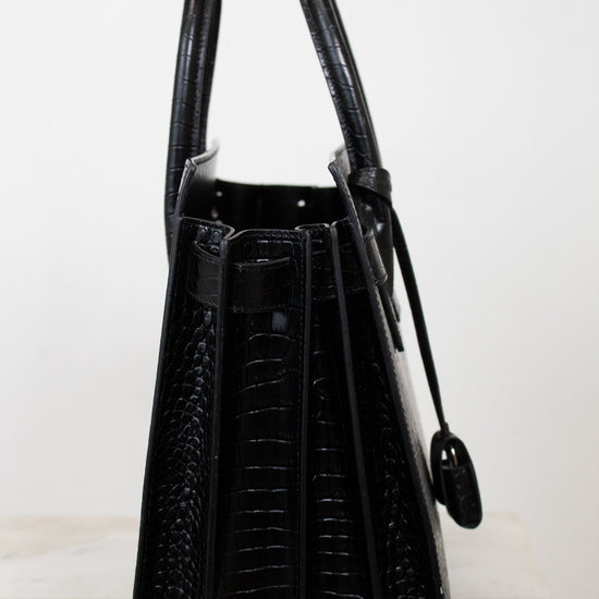 A black crocodile-embossed leather handbag shown from the side, featuring dual top handles and a hanging clochette, set against a plain white background.