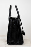 A black crocodile-embossed leather handbag shown from the side, featuring dual top handles and a hanging clochette, set against a plain white background.
