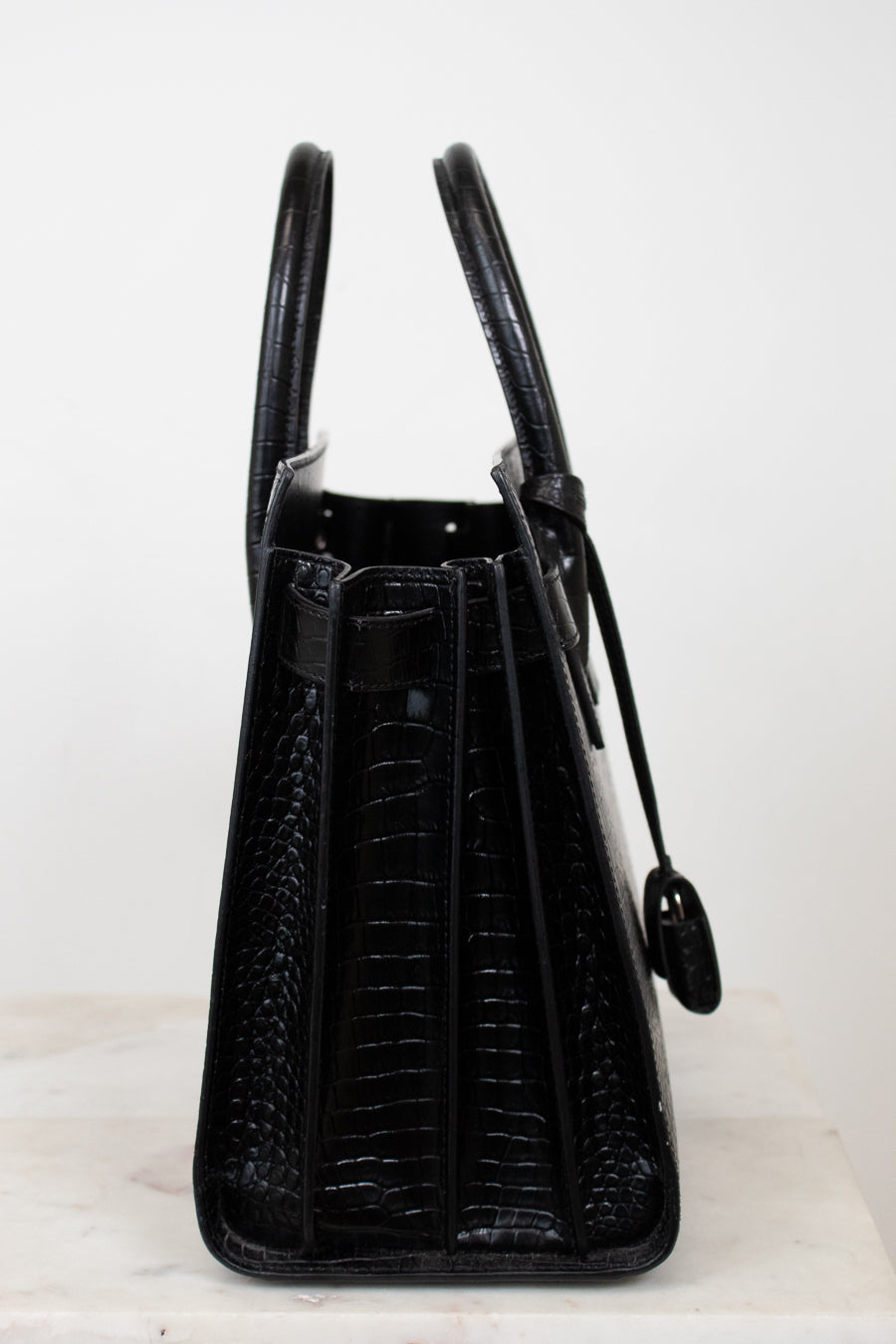 A black crocodile-embossed leather handbag shown from the side, featuring dual top handles and a hanging clochette, set against a plain white background.