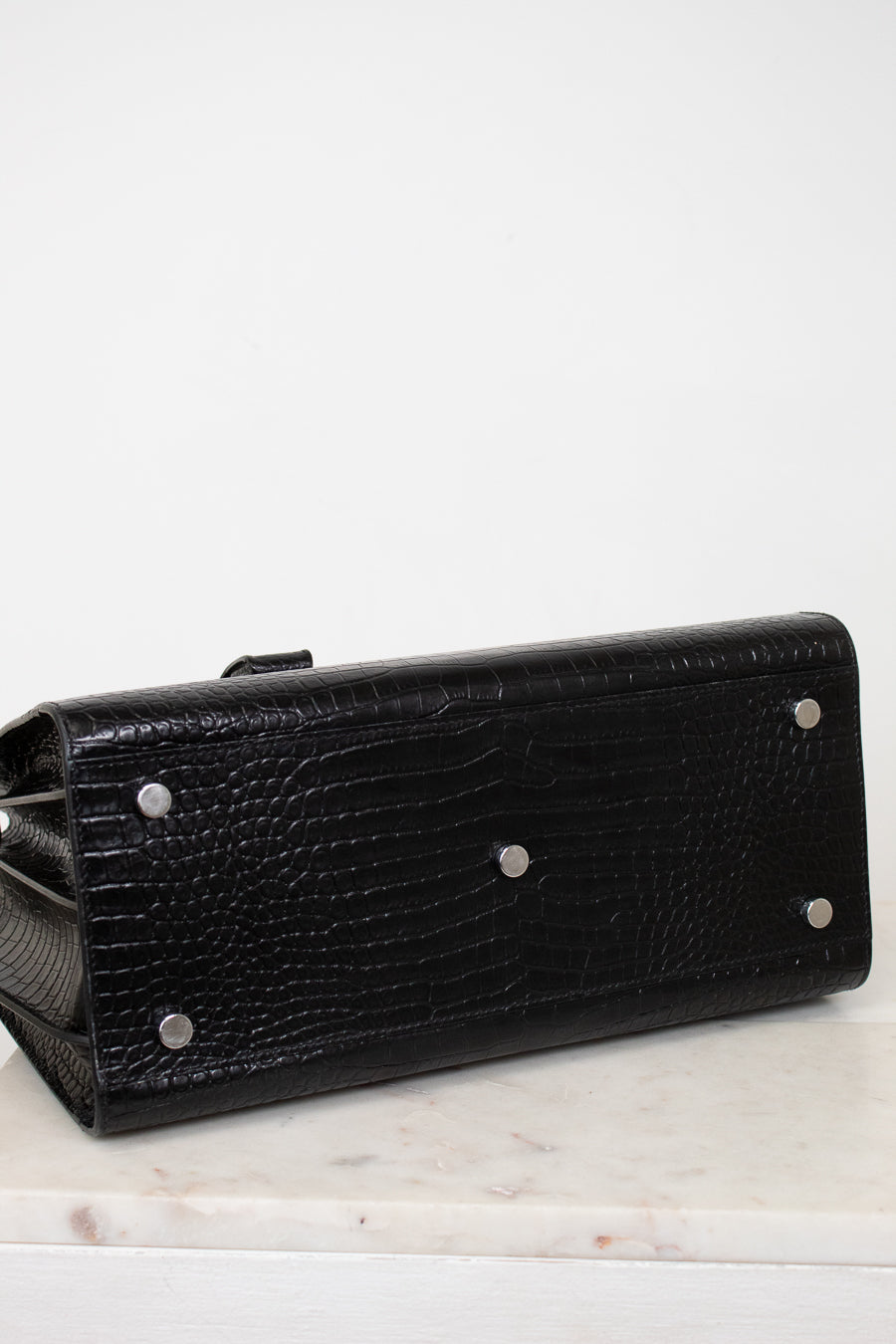 A black textured leather clutch purse with silver-tone metal studs, shown from a side angle on a light marble surface against a white background.