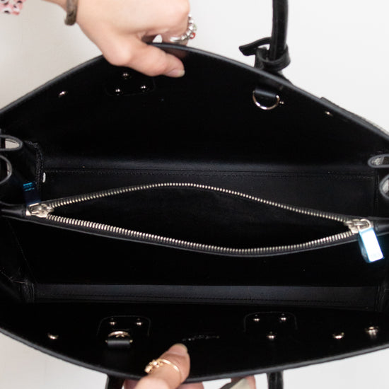 Close-up of a black leather handbag interior with a top zipper, held open by two hands against a plain light background.