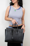 A woman wearing a light gray ribbed tank top and black pants holds a large black crocodile-embossed leather handbag with top handles and a front flap detail against a plain light background.
