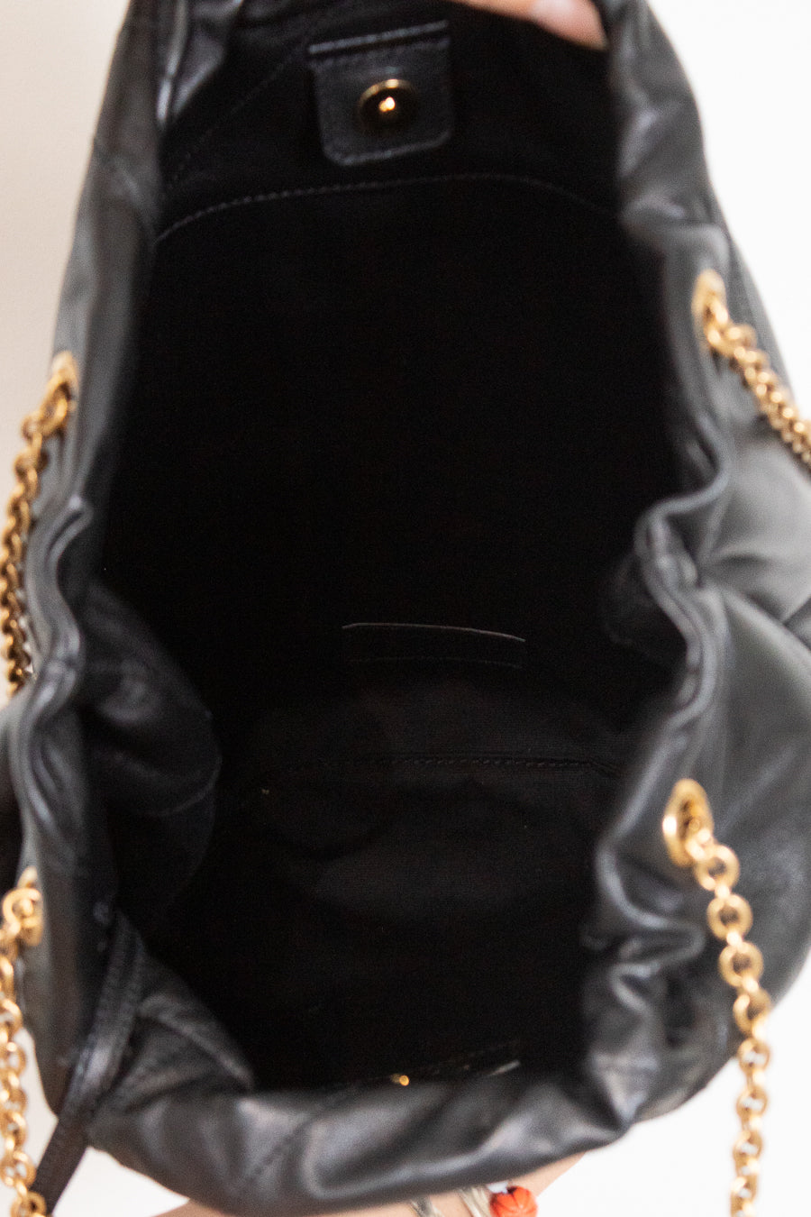 Black leather bucket bag shown from the inside, featuring a black fabric lining, a gold-tone snap button closure, and gold chain straps visible at the sides.