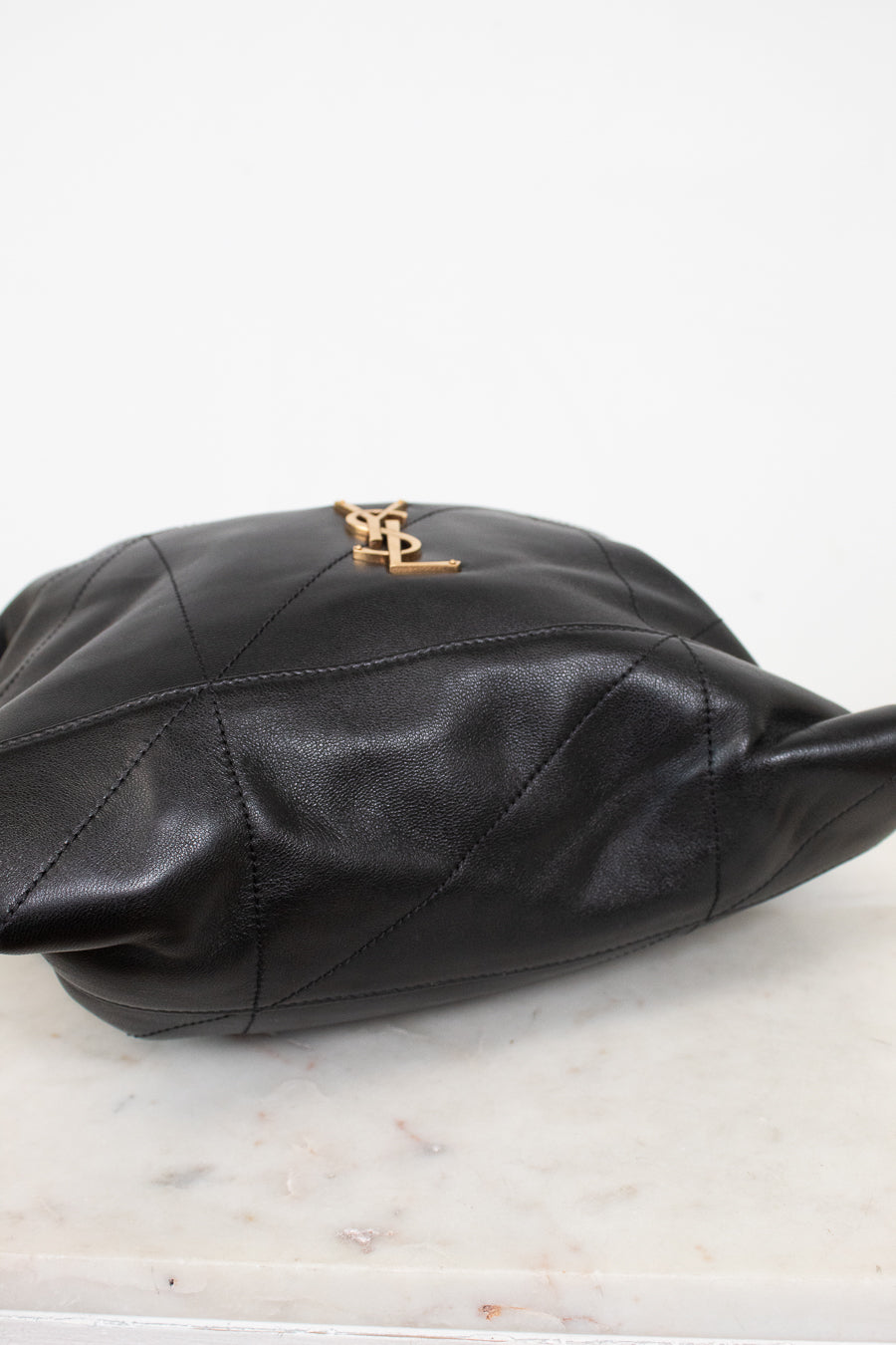 A black leather shoulder bag with a gold-tone YSL logo clasp, resting on a white marble surface against a white background.