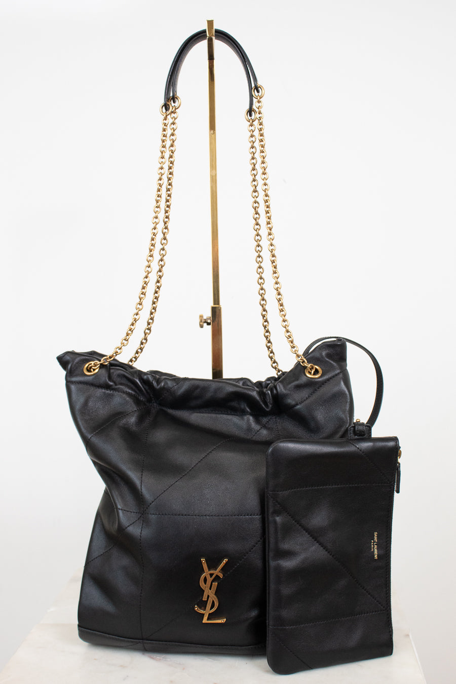 A black leather Saint Laurent shoulder bag with gold-tone chain straps and a matching black leather pouch, displayed on a white background with a stand.