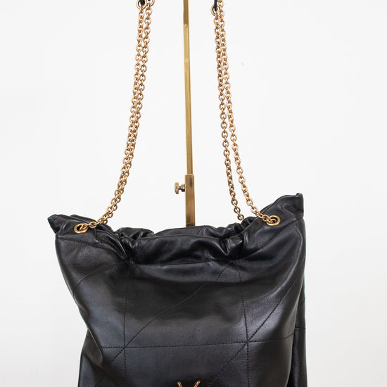 A black leather shoulder bag with a gold-tone YSL hardware logo, featuring a quilted pattern, a drawstring top, and a gold chain strap, displayed on a stand against a white background.