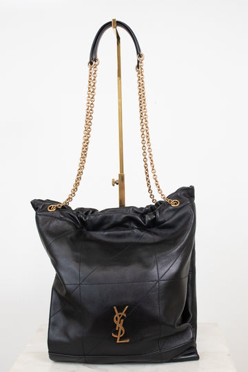 A black leather shoulder bag with a gold-tone YSL hardware logo, featuring a quilted pattern, a drawstring top, and a gold chain strap, displayed on a stand against a white background.