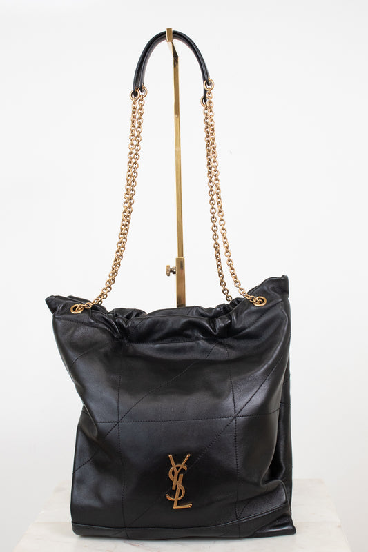 A black leather shoulder bag with a gold-tone YSL hardware logo, featuring a quilted pattern, a drawstring top, and a gold chain strap, displayed on a stand against a white background.