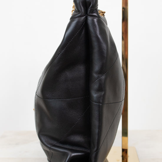 A black leather slouchy shoulder bag with gold-tone chain straps, displayed on a gold stand against a white background.