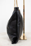 A black leather slouchy shoulder bag with gold-tone chain straps, displayed on a gold stand against a white background.