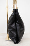 A black leather slouchy bucket bag with gold chain straps, displayed on a gold stand against a white background.