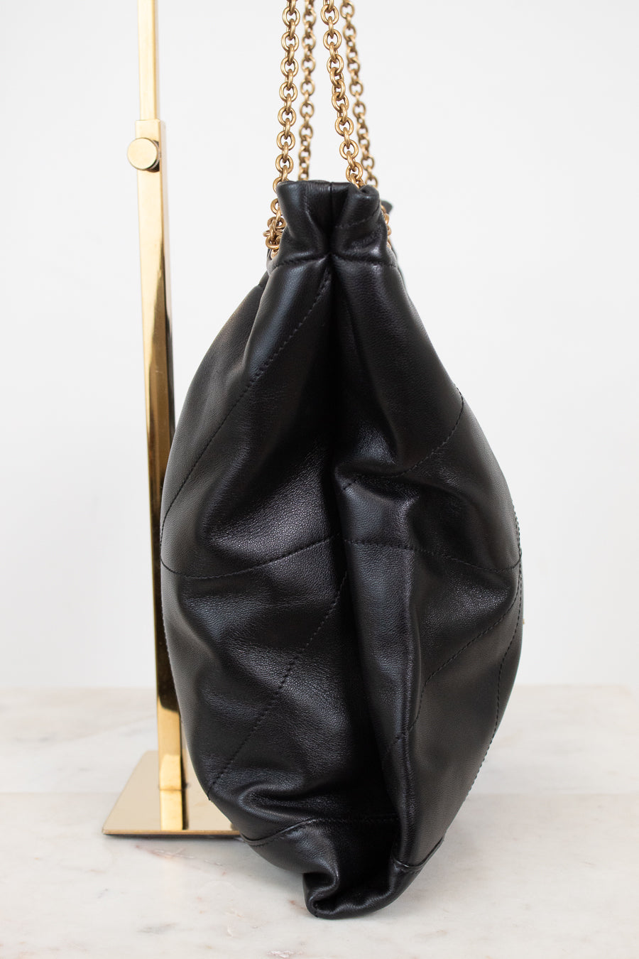 A black leather slouchy bucket bag with gold chain straps, displayed on a gold stand against a white background.