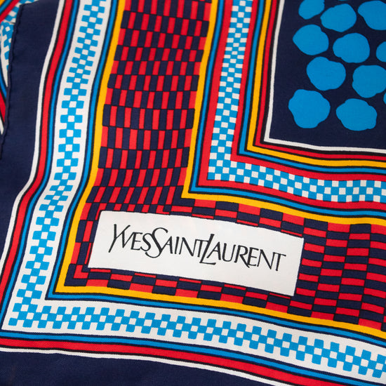A close-up of a colorful Yves Saint Laurent scarf featuring a geometric pattern with navy blue, red, light blue, yellow, and white, complete with the brand's signature white label.