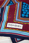 A close-up of a colorful Yves Saint Laurent scarf featuring a geometric pattern with navy blue, red, light blue, yellow, and white, complete with the brand's signature white label.