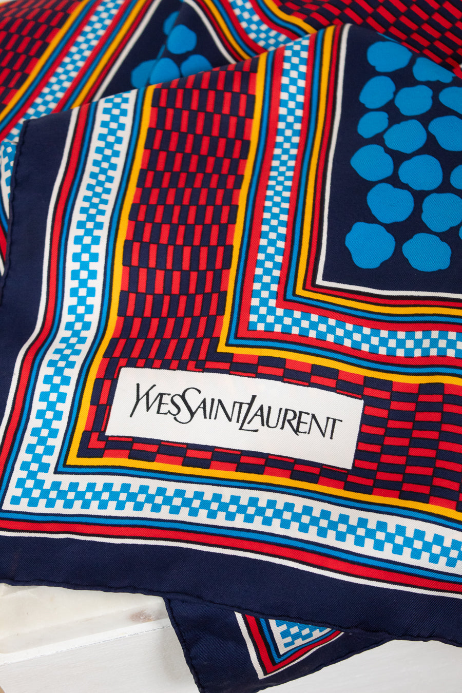 A close-up of a colorful Yves Saint Laurent scarf featuring a geometric pattern with navy blue, red, light blue, yellow, and white, complete with the brand's signature white label.