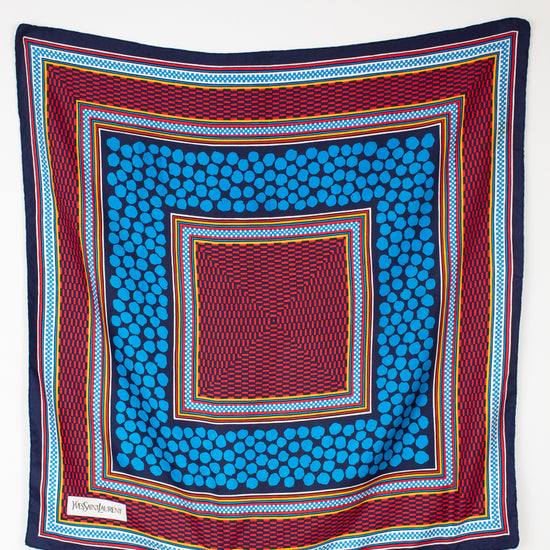 A square scarf with a vibrant geometric pattern, featuring concentric squares of blue, red, navy, and yellow on a plain white background.