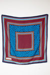 A square scarf with a vibrant geometric pattern, featuring concentric squares of blue, red, navy, and yellow on a plain white background.