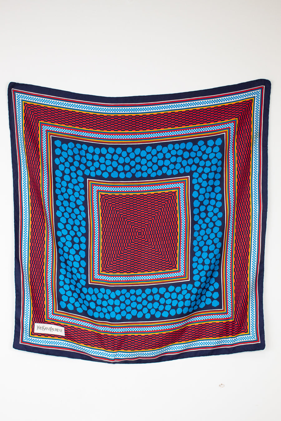 A square scarf with a vibrant geometric pattern, featuring concentric squares of blue, red, navy, and yellow on a plain white background.