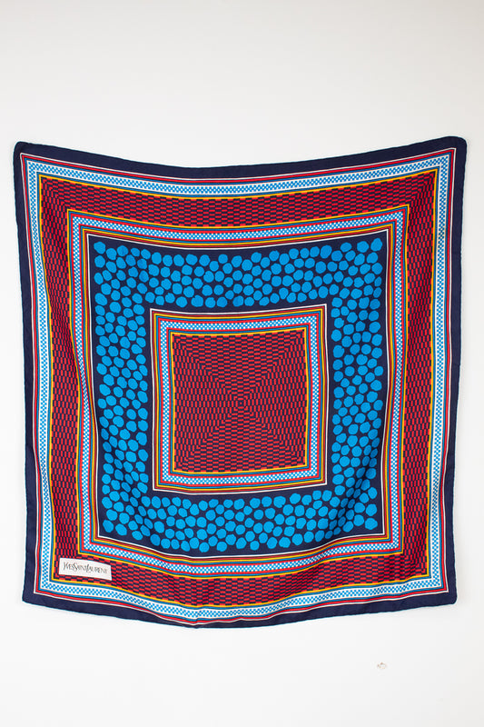 A square scarf with a vibrant geometric pattern, featuring concentric squares of blue, red, navy, and yellow on a plain white background.