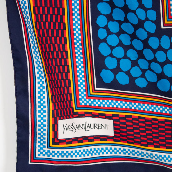 A close-up of a Yves Saint Laurent scarf showing its corner, featuring a navy blue base with a blue polka-dot pattern, a red and blue geometric checkered border, and a white brand label.