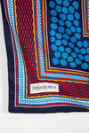 A close-up of a Yves Saint Laurent scarf showing its corner, featuring a navy blue base with a blue polka-dot pattern, a red and blue geometric checkered border, and a white brand label.