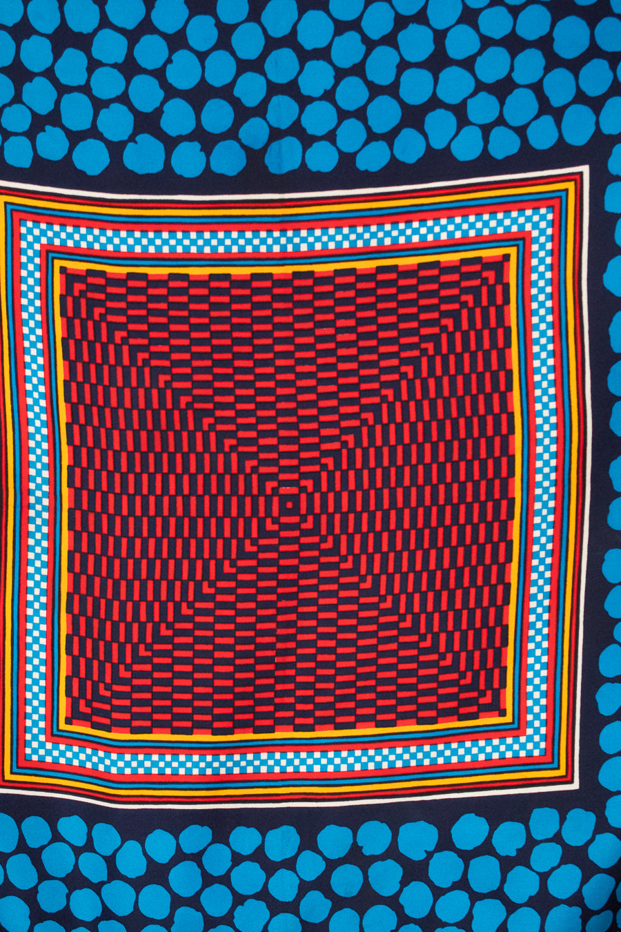 A square scarf with a bold geometric pattern, featuring a central red, black, and blue abstract checkerboard-like grid surrounded by concentric multicolor borders, with a dark navy background patterned with bright blue dots at the top and bottom.