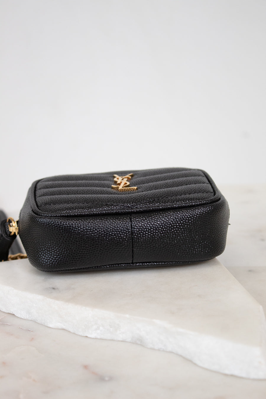 A black pebbled leather pouch with a gold-tone YSL logo on the front, resting on a white marble slab against a light gray background.