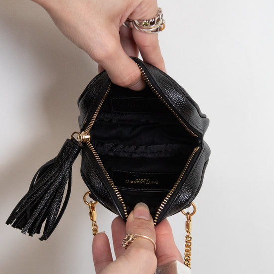 A close-up of a black textured leather mini pouch bag with gold-tone hardware, held open by two hands to show the interior. The bag features a black leather tassel zipper pull and a gold chain strap
