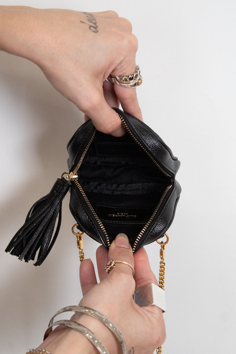 A close-up of a black textured leather mini pouch bag with gold-tone hardware, held open by two hands to show the interior. The bag features a black leather tassel zipper pull and a gold chain strap