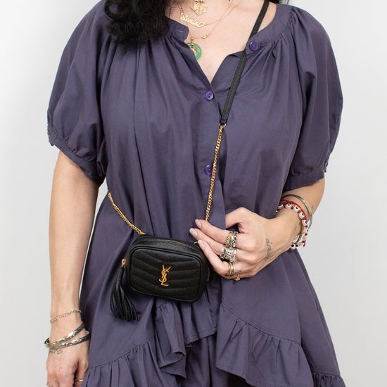 A woman wearing a short-sleeve, ruffled mini dress in a muted purple shade, styled with a black quilted crossbody bag featuring a gold chain strap and YSL monogram.