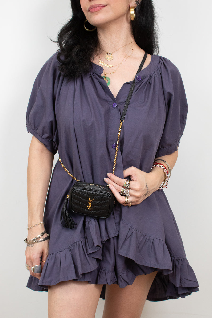 A woman wearing a short-sleeve, ruffled mini dress in a muted purple shade, styled with a black quilted crossbody bag featuring a gold chain strap and YSL monogram.