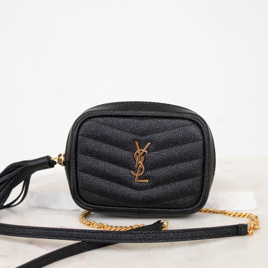 A black quilted leather crossbody bag with a gold-tone YSL monogram logo and a gold chain strap, resting on a white marble surface against a white background.