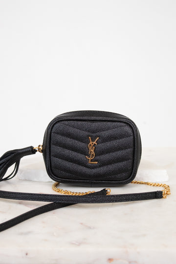 A black quilted leather crossbody bag with a gold-tone YSL monogram logo and a gold chain strap, resting on a white marble surface against a white background.