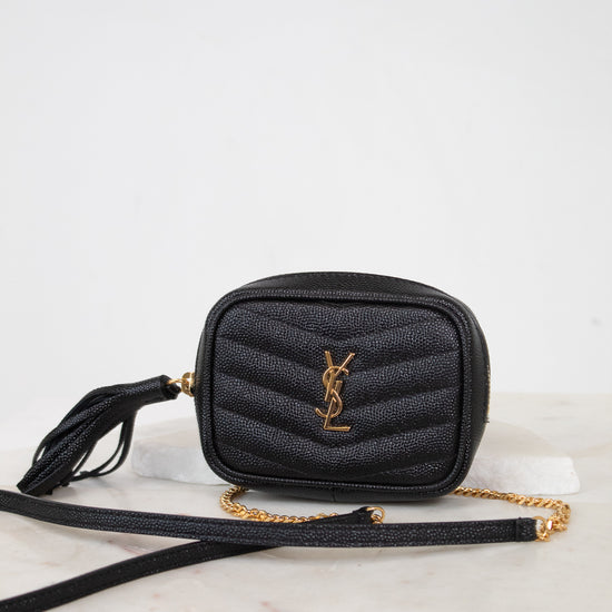 A black quilted leather crossbody bag with a gold-tone YSL monogram logo and a gold chain strap, resting on a white marble surface against a white background.
