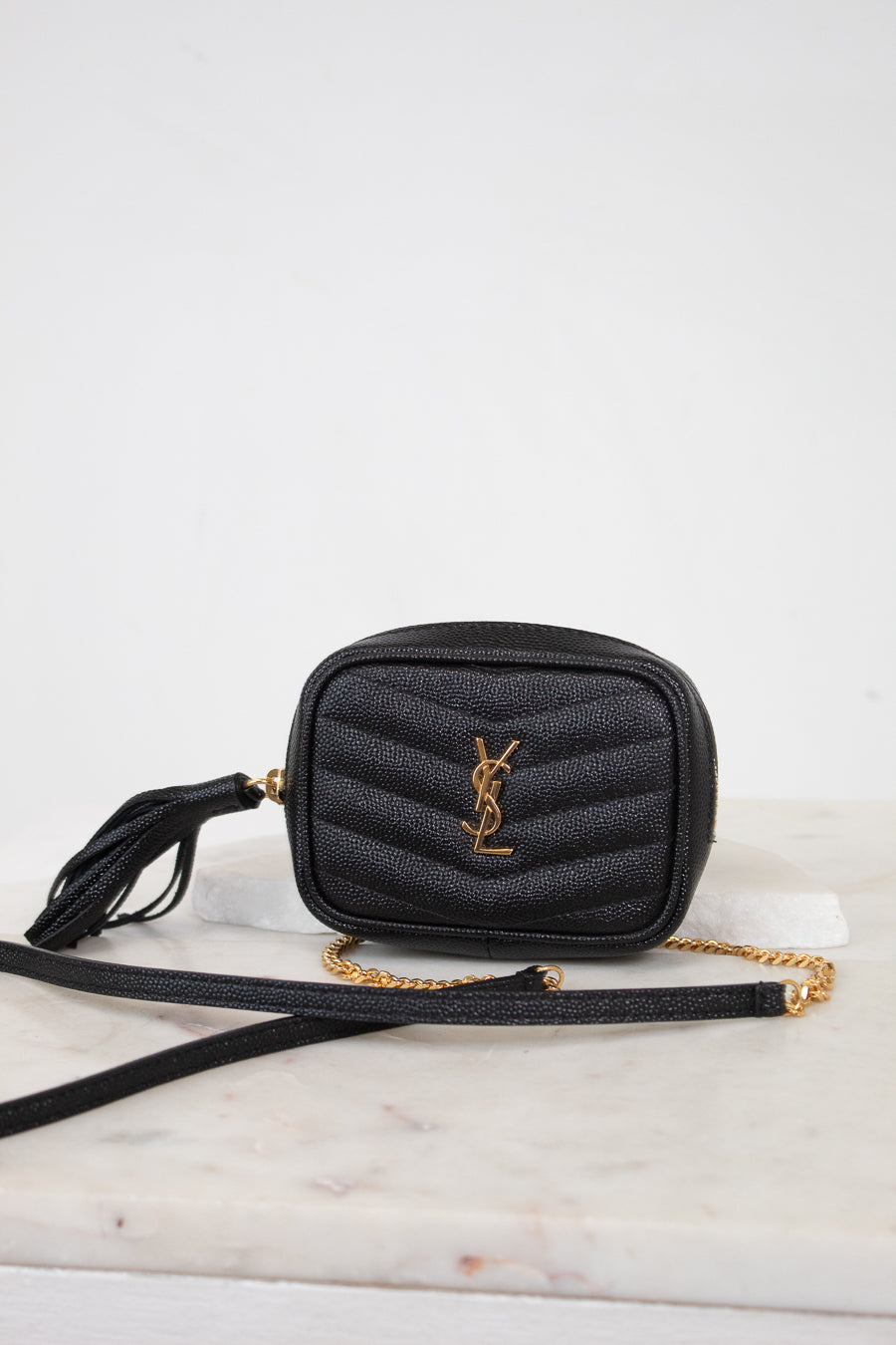A black quilted leather crossbody bag with a gold-tone YSL monogram logo and a gold chain strap, resting on a white marble surface against a white background.