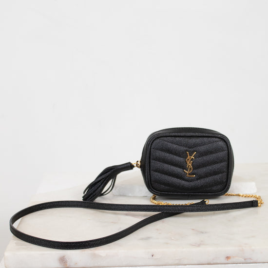 A black quilted leather crossbody bag with a gold-tone YSL monogram logo on the front, resting on a white marble slab against a white background.