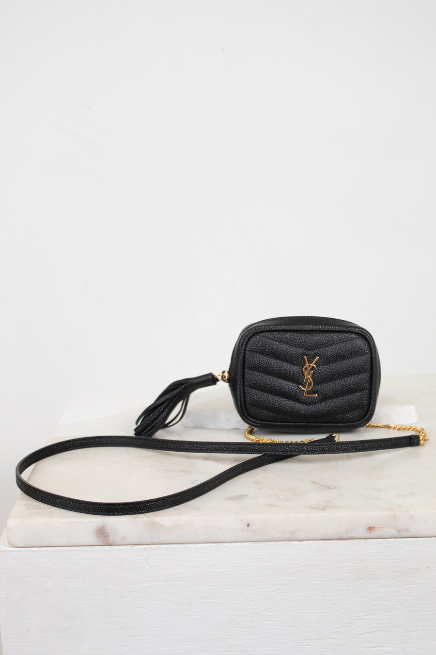 A black quilted leather crossbody bag with a gold-tone YSL monogram logo on the front, resting on a white marble slab against a white background.