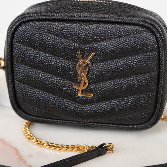 A black textured leather camera bag with a gold-tone YSL monogram logo on the front, resting on a light marble surface with a gold chain strap visible.