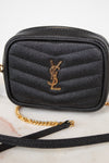 A black textured leather camera bag with a gold-tone YSL monogram logo on the front, resting on a light marble surface with a gold chain strap visible.