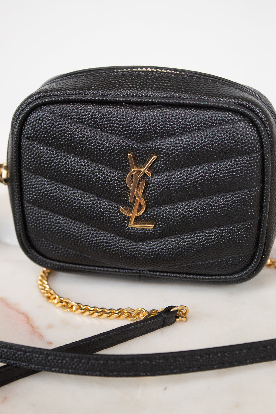 A black textured leather camera bag with a gold-tone YSL monogram logo on the front, resting on a light marble surface with a gold chain strap visible.