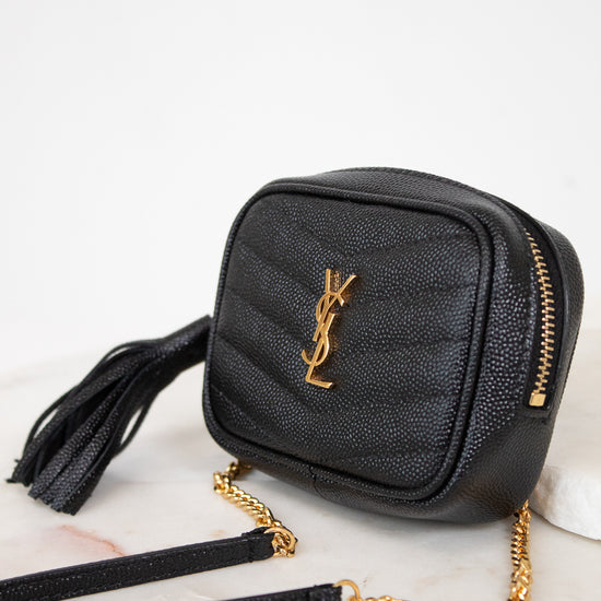 A black textured leather crossbody bag with a gold-tone YSL monogram logo, gold zipper, and a gold chain strap, resting on a white marble surface.
