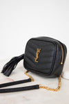 A black textured leather crossbody bag with a gold-tone YSL monogram logo, gold zipper, and a gold chain strap, resting on a white marble surface.