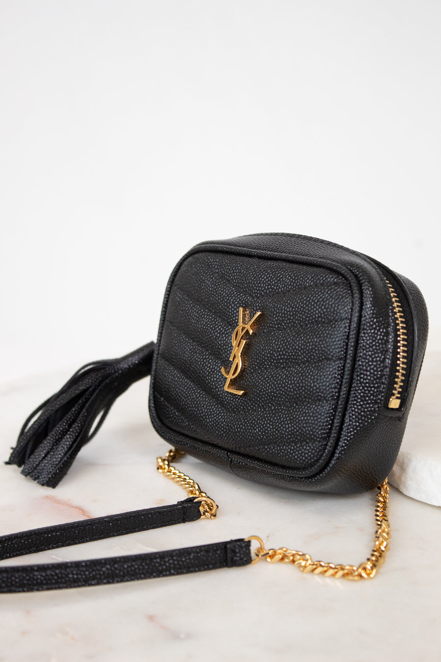 A black textured leather crossbody bag with a gold-tone YSL monogram logo, gold zipper, and a gold chain strap, resting on a white marble surface.