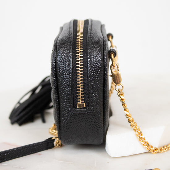 A black pebbled leather crossbody camera bag with gold-tone hardware, a gold zipper, a black tassel, and a gold chain strap, shown on a white marble surface against a white background.