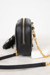 A black pebbled leather crossbody camera bag with gold-tone hardware, a gold zipper, a black tassel, and a gold chain strap, shown on a white marble surface against a white background.